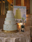 This blue fondant-covered cake is piped with royal icing and painted with gold luster dust. Oval rococo frames of foil-covered paper are attached to the tiers with royal icing as are the ribbon bows. Metallic lace trims the tiers.