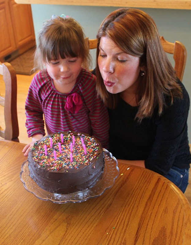 Lexi and daughter blowing out candles of a birthday version of Hershey’s Perfect Chocolate Cake sitting on a glass cake plate