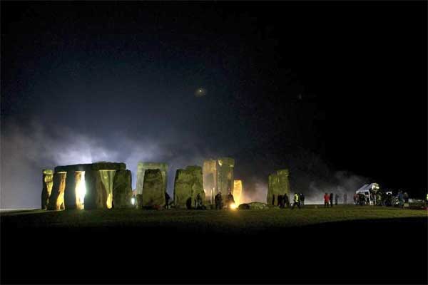 Stonehenge by night, with a single bright star in the sky and mist rising from the brightly floodlit stones. There are people around the stones, some in brightly coloured jackets.  To the right of the image is a large white van with people gathered around it and some unclear shapes which may be other vehicles.