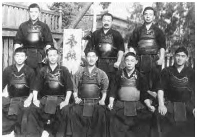 Teachers and future teachers of the Hokubei Butokukai, Japan, ca. 1936. In the back row are Yamamoto (1-dan), Nakamura Sensei (6-dan), and Hirano (5-dan). The front row includes Hara (2-dan), Muruyama (4-dan), Fujii Sensi (4-dan), and Imada (2-dan). Although partially blocked, the sign appears to read "dedication meeting." 
