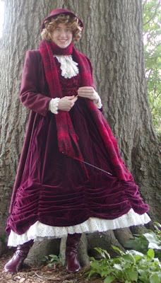 Young woman wearing mid-calf length Victorian gown in burgundy, somewhat ruched up at the bottom to expose a lacy petticoat. She wears burgundy lace-up boots, a white lace jabot, and is knitting an enormous burgundy scarf that is looped around her neck. She is smiling at the viewer, has curly brown hair, and is wearing a dainty burgundy hat.