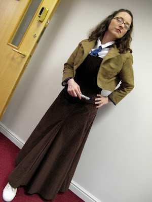 Joanna Mead wears a Victorian-inspired ankle-length skirt and matching over-the-bust bodice, both in pinstriped brown velveteen, as she looks haughtily at the viewer. She is wearing a white mens' dress shirt under the bodice; the collar and blue necktie are visible above the bodice. She is wearing a brown bolero jacket of the same color, material, and lapel cut of David Tennant's coat. In her right hand is a sonic screwdriver toy.