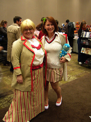 Two young women in similar, but distinct, Fifth Doctor variant costumes. They are facing the viewer. The one on the left has a blouse with a red ruffle sewn on, in imitation of the red chevron knitted into Peter Davison's sweater, and is wearing an ankle-length striped skirt. On her left (viewer's right) is a woman wearing a sweater and a much shorter (mid-thigh length) striped skirt.