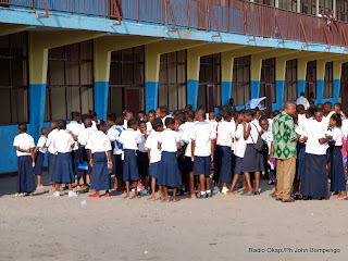 En avant plan de droite à gauche en chemise verte un enseignant en conversation avec des élèves habillés en bleu blanc devant le bâtiment d'une école à Kinshasa. Radio Okapi/ Ph John Bompengo