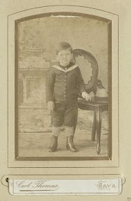 Portret van een jongetje in matrozenpak bij een stoel tegen een achtergronddoek