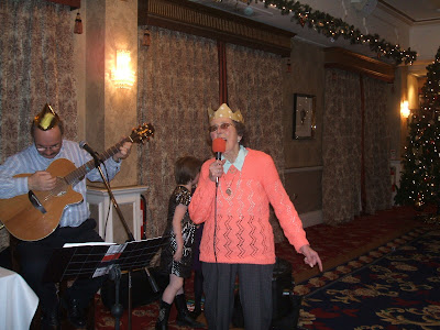 An old woman obviously enjoying singing along with a band in a big Hotel room. A Christmas tree is visible in the corner.