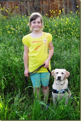 My niece Sara standing in a field of yellow mustard flowers with Reyna sitting next to her in her puppy coat.  Sara has a very cute smile on her face.