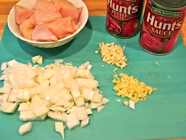 prepped ingredients - cubed chicken in bowl, chopped onion, garlic and ginger on cutting board with can on tomato sauce and paste in background. 