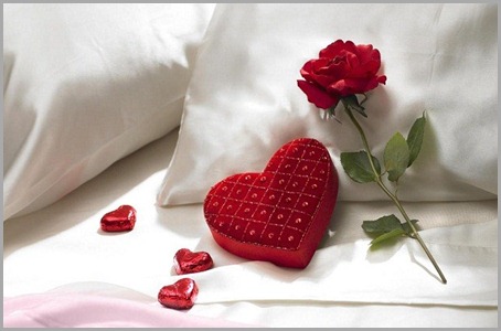 Valentine's Day romantic red rose and heart shaped candy box with red heart shaped chocolate candy on pillow of bed.