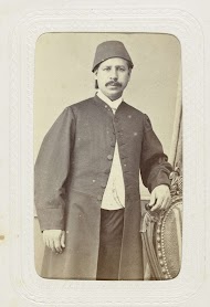 Studioportret van een man met snor in lange jas met op zijn hoofd een fez.