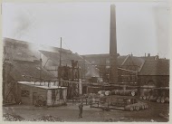 Fabrieksterrein met gebouwen, een schoorsteen, houten vaten en een werknemer