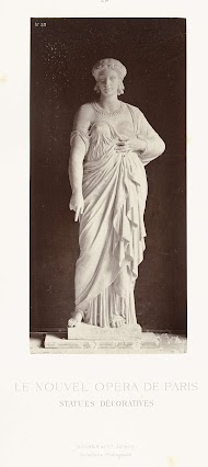 Marmeren beeld van een vrouw in een gewaad met een ontblote borst, de wijsvinger aan haar rechterhand wijst naar beneden.